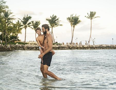 Two people are in shallow water at a beach; one is carrying the other on their back. Palm trees and a cloudy sky are in the background.