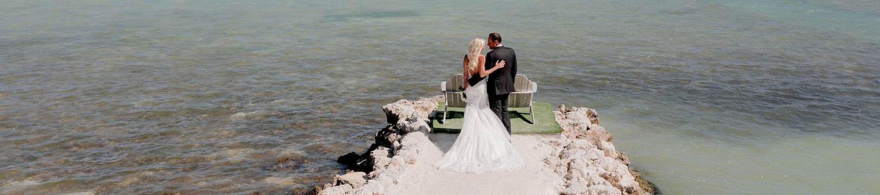 A bride and groom stand on a rocky pier extending into the turquoise ocean, embracing each other on their wedding day.