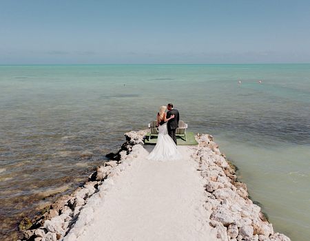 A bride and groom stand on a rocky pier extending into the turquoise ocean, embracing each other on their wedding day.