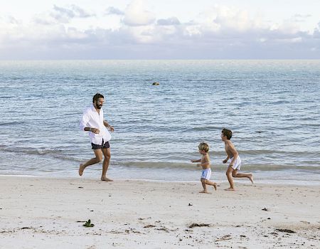 A man and two children are running and playing on a sandy beach by the ocean under a partly cloudy sky.