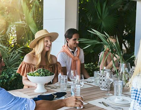 A group of people sitting around an outdoor table enjoying a meal, with greenery in the background and sunlight filtering through.