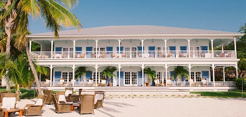 A two-story building with balconies, surrounded by palm trees, and outdoor seating on sandy ground, under a clear blue sky.