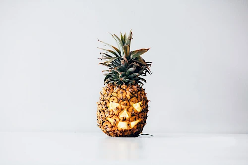 A pineapple is carved with a face, resembling a jack-o'-lantern, and lit from within, placed on a white surface.