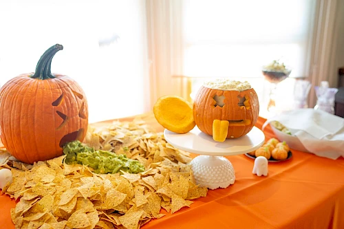 Two carved pumpkins on a table with chips and guacamole arranged creatively, one looks like it's vomiting, creating a fun Halloween theme.