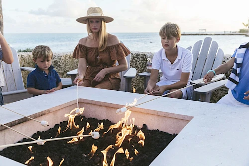 People are gathered around a fire pit by the sea, roasting marshmallows on sticks while sitting on Adirondack chairs.