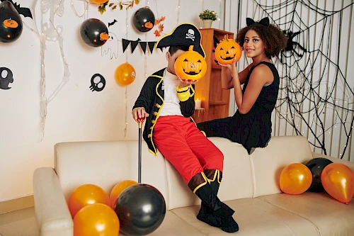 Two kids in Halloween costumes hold pumpkin props on a decorated sofa with balloons, cobwebs, and hanging decorations.