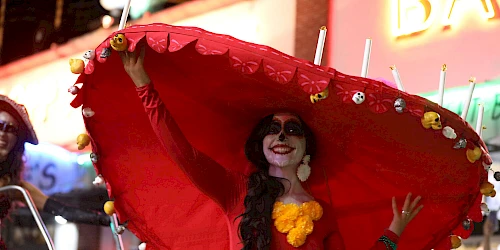 A person in festive Day of the Dead attire with face paint and a large red hat decorated with candles and skulls, posing with a smile.