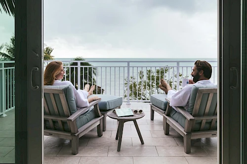 A woman and a man in robes relax on a balcony, sitting in chairs, enjoying drinks, with a scenic ocean view in the background.