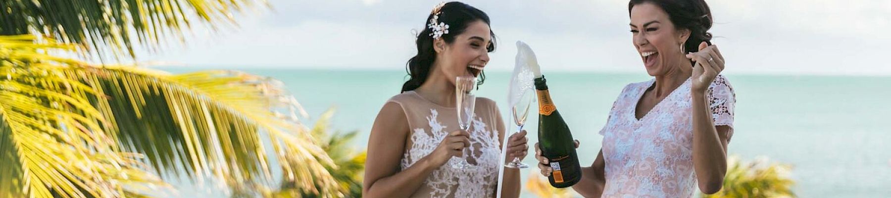 Two women in dresses are on a balcony with a scenic view, celebrating and opening a champagne bottle.