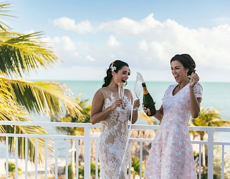 Two women in dresses are on a balcony with a scenic view, celebrating and opening a champagne bottle.