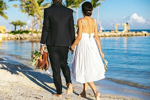 A couple walks barefoot along a beach, carrying shoes and a bouquet, with palm trees and the ocean in the background.