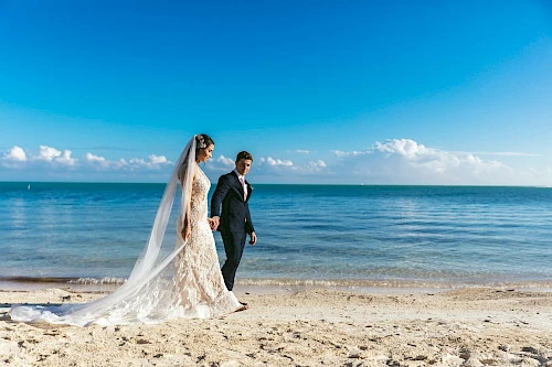 A bride and groom walk on a beach, with the sea and a clear blue sky in the background.