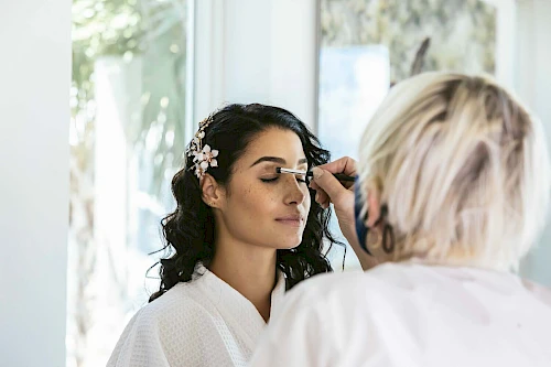 A person is applying makeup to another person's face while they sit in a well-lit room wearing a white robe and floral hair accessory.