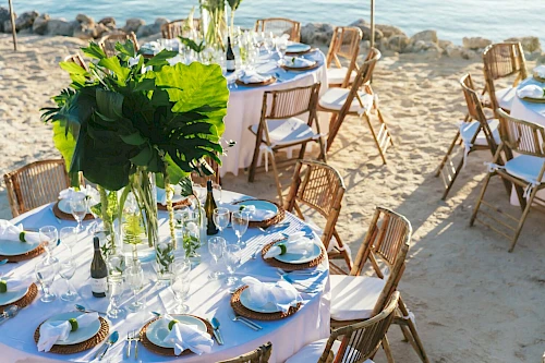 Beachfront dining setup with round tables, white linens, greenery centerpieces, and wooden chairs on sand, near the water.