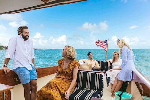 A group of people enjoy a sunny day on a boat, chatting and relaxing with clear blue skies and ocean in the background, an American flag visible.