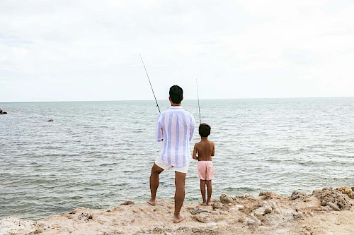 Two people fishing on a rocky shoreline with the ocean in the background, both holding fishing rods, facing away from the camera.