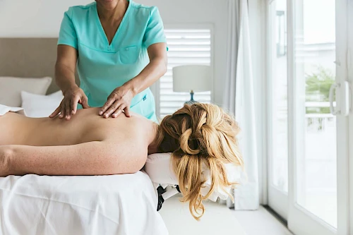 A person is receiving a back massage from a massage therapist in a bright room with large windows.