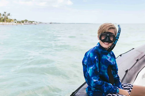 A person with a snorkel and mask sits on the edge of a boat in a calm, blue ocean with a distant shoreline and cloudy sky in the background.