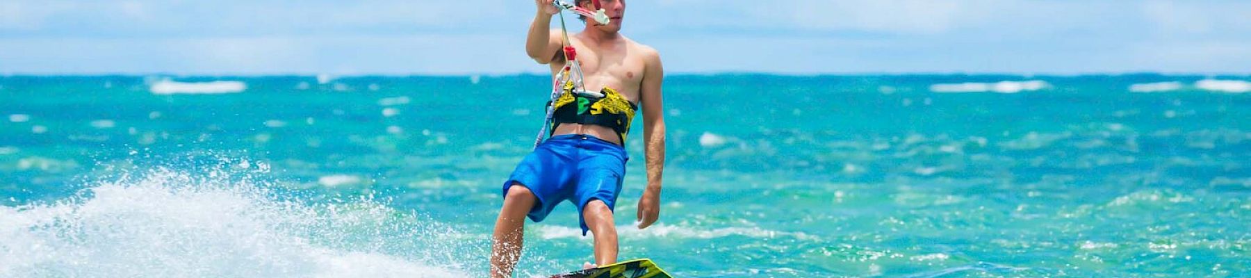 A person is kiteboarding on clear, blue waters under a bright sky, creating splashes as they glide across the sea.