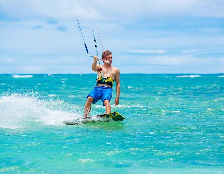 A person is kiteboarding on clear, blue waters under a bright sky, creating splashes as they glide across the sea.