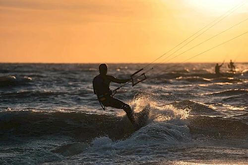 A silhouette of a person kite surfing on the ocean at sunset, with waves visible and another surfer in the background under the golden sky.