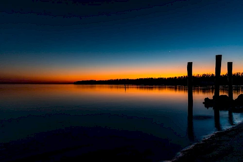 A serene sunset over calm waters with silhouettes of poles and distant trees against an orange and blue gradient sky.