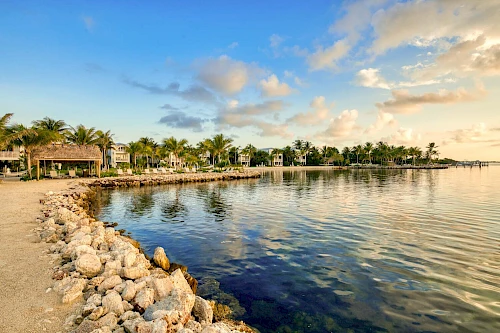 A serene coastal scene with calm waters, a rocky shoreline, lush palm trees, and a partly cloudy sky during sunset or sunrise.