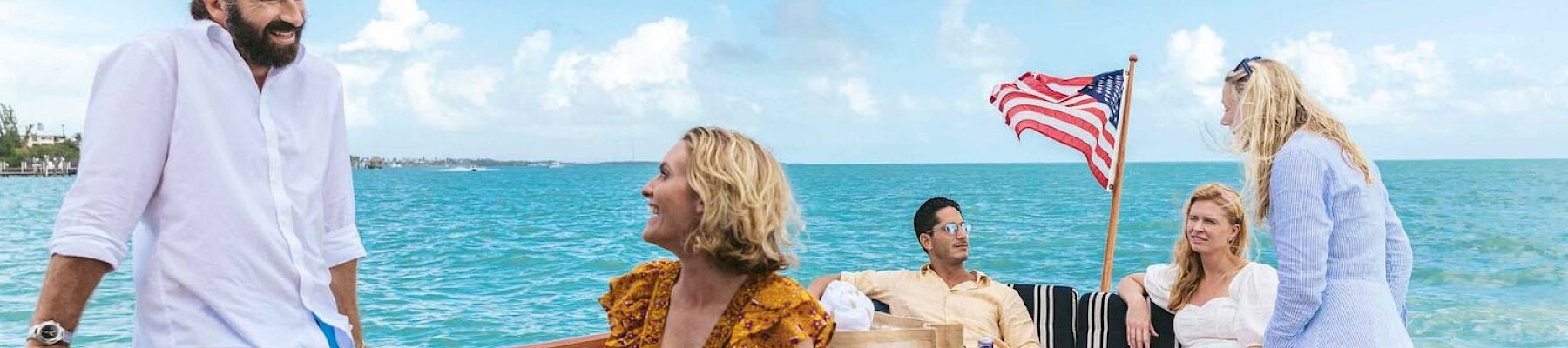 A group of people is enjoying a sunny day on a boat with an American flag, relaxed and conversing with a bright blue sky in the background.