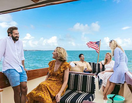 A group of people is enjoying a sunny day on a boat with an American flag, relaxed and conversing with a bright blue sky in the background.