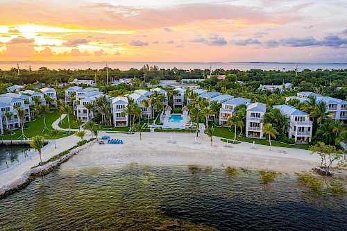 Aerial view of a beachfront resort with modern buildings, a pool, palm trees, and a sandy shoreline at sunset over the ocean.