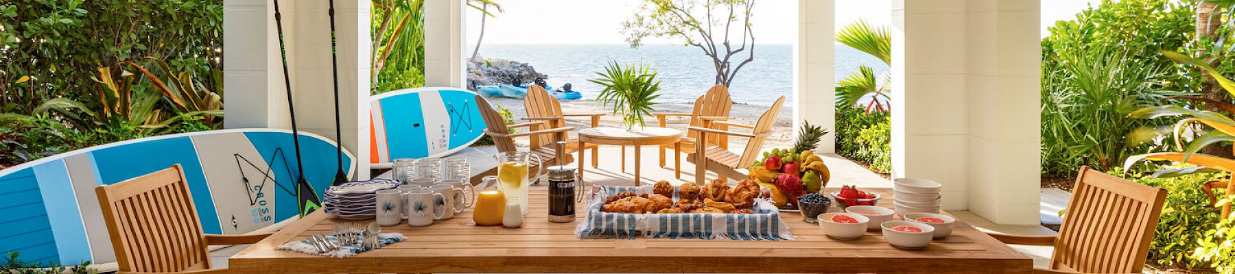 An outdoor covered patio with a wooden dining set, assorted breakfast items, and surfboards, overlooking a garden and the ocean in the background.