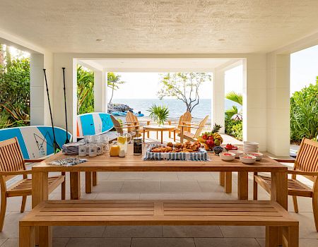 An outdoor covered patio with a wooden dining set, assorted breakfast items, and surfboards, overlooking a garden and the ocean in the background.