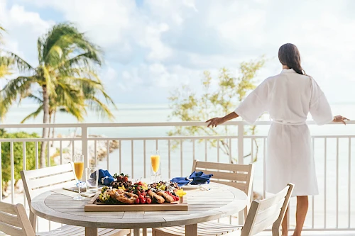 A person in a robe overlooks the ocean from a balcony with a table set for breakfast, including fruit and orange juice.