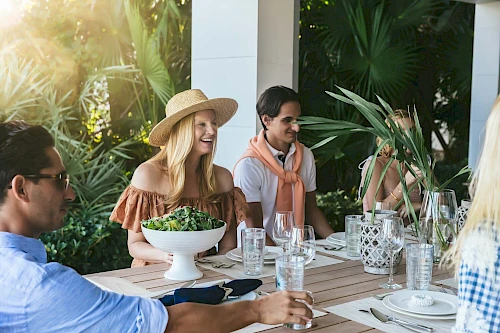 People are enjoying an outdoor meal at a table surrounded by plants, with glasses, plates, and a salad bowl set out.