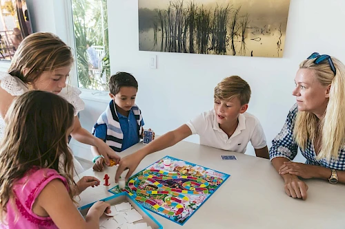 A group of people are playing a colorful board game at a table, watched by a woman in a blue checkered shirt.