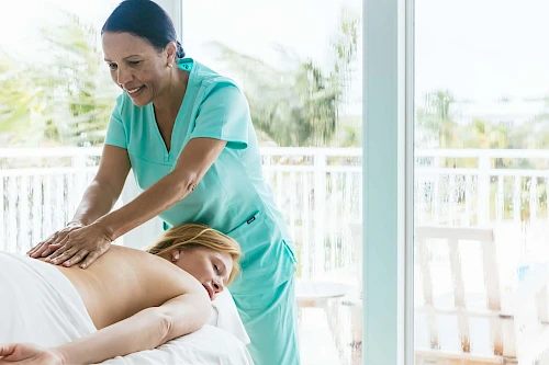 A person in a spa setting is receiving a massage from a therapist in a bright room with large windows.