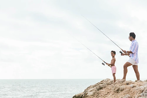 Two people are fishing on a rocky shoreline, one adult and one child, both holding fishing rods and looking out at the ocean, under a cloudy sky.