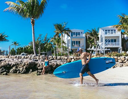 A man carrying a paddleboard is running towards the water on a sunny beach with palm trees and modern beachfront houses in the background.