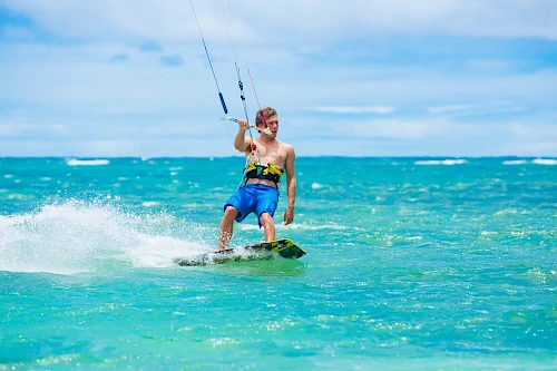 A person is kite surfing on a clear, blue ocean under a light blue sky with some clouds, wearing blue shorts and a life vest.