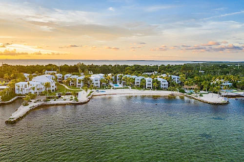 A coastal residential area with modern white homes along a calm bay, rocky shoreline, and a warm sunset casting golden hues over the water.