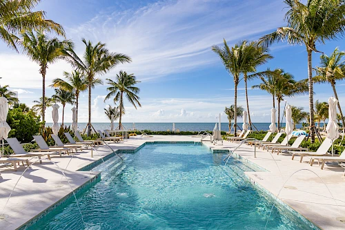 A luxurious poolside area with palm trees, sun loungers, and an ocean view under a clear blue sky.