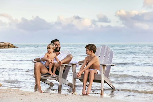 A person is sitting in a chair at the beach, holding a child, with another child sitting in a separate chair nearby, enjoying the ocean view.