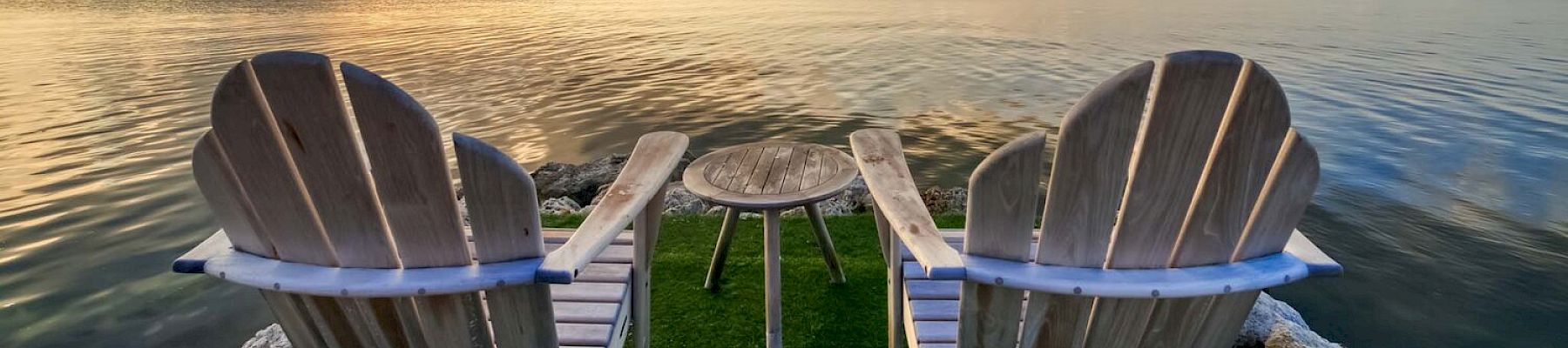 Two wooden Adirondack chairs face a calm sea at sunset, with a small wooden table between them, under a stunning, colorful sky.