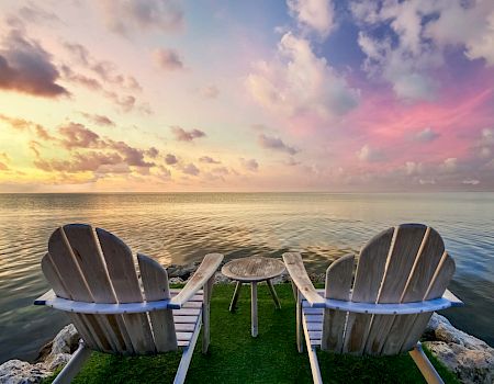 Two wooden Adirondack chairs face a calm sea at sunset, with a small wooden table between them, under a stunning, colorful sky.