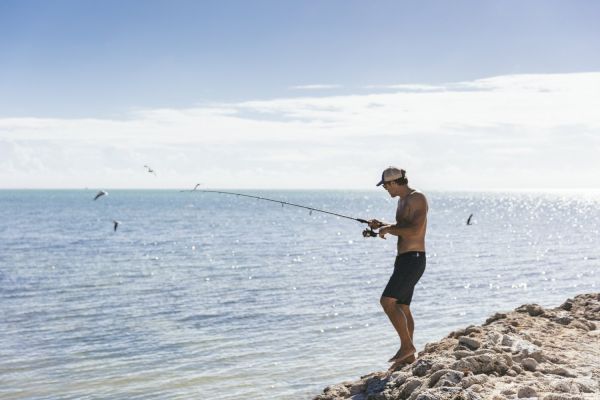 A person is standing on rocky terrain near the sea, fishing with a fishing rod, and wearing shorts and a cap.