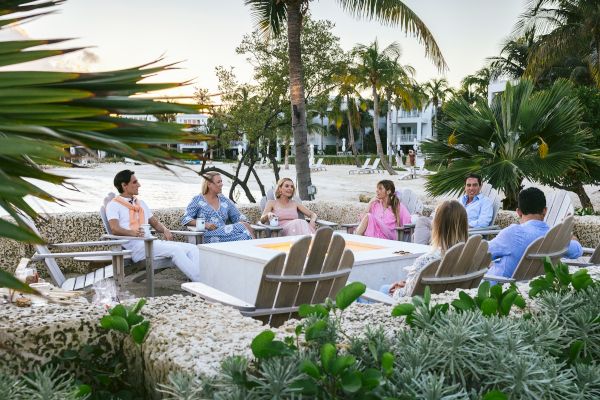 A group of people is sitting and socializing outdoors on a beach, surrounded by palm trees and greenery, with some buildings in the background.