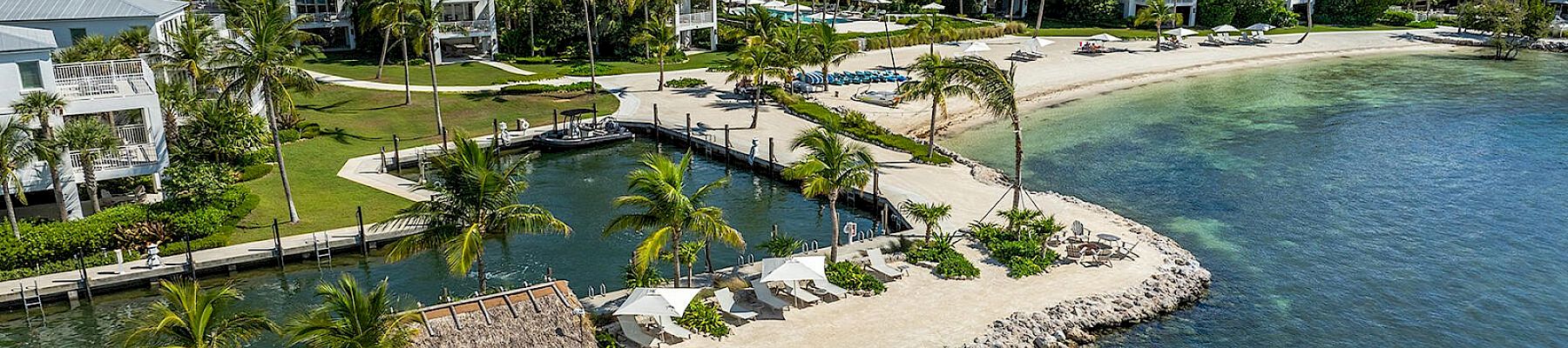 A waterfront resort area with palm trees, white buildings, a small lagoon, and a sandy beach bordering clear green-blue water.