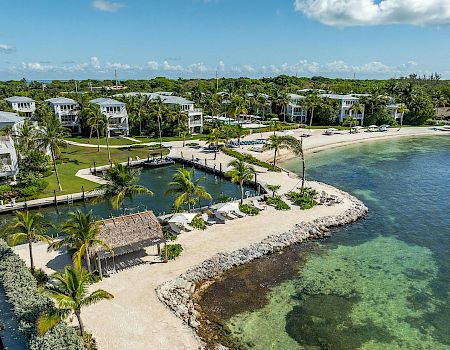 A waterfront resort area with palm trees, white buildings, a small lagoon, and a sandy beach bordering clear green-blue water.