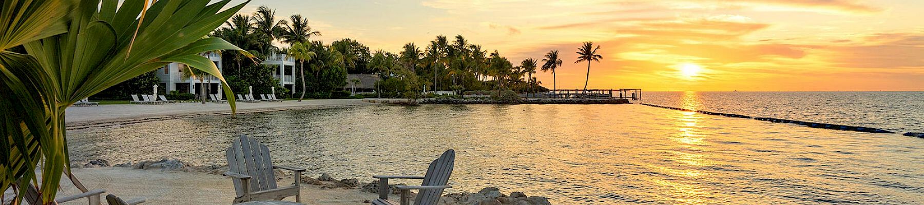 A serene beach setting with four wooden chairs around a fire pit, overlooking a tranquil sunset on the water, surrounded by palm trees.