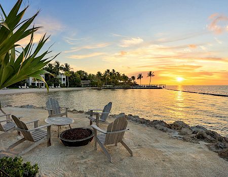 A serene beach setting with four wooden chairs around a fire pit, overlooking a tranquil sunset on the water, surrounded by palm trees.